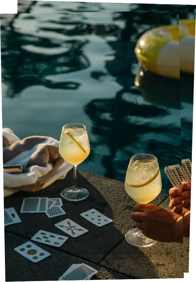 Vertical postcard of a close-up of a card game on the edge of a harbor by the water. There are two glasses with Allora drink in them, one on the ground and the other held by someone's hand.