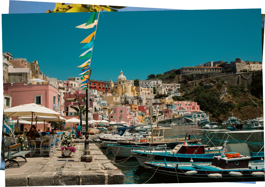 Horizontal Postcard of Procida's harbor with boats on the right and waterfront restaurants on the left. There are flags waving in the wind and the colourful pastel building of Procida in the background.