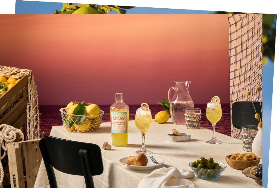 Horizontal postcard of a table with beige tablecloth and two black chairs on either side. The table has an open bottle of Allora on it, two glasses with Allora drink in them, a basket of lemons and other small items such as olives.
