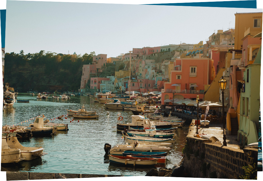 Horizontal postcard showing the colourful pastel buildings of the Procida seafront facing the harbor with boats parked in it