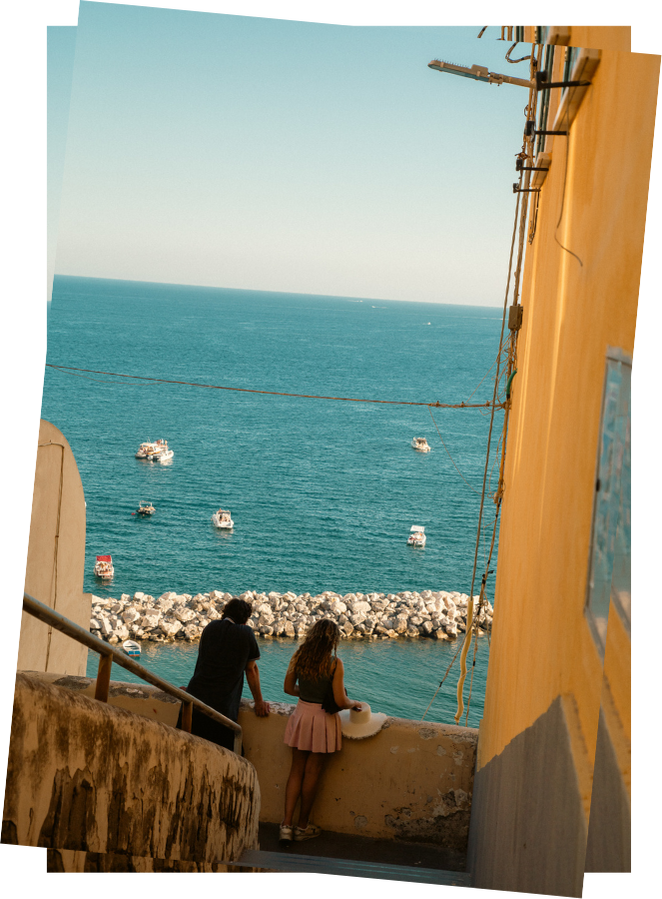 Vertical Postcard of two people in the streets of Procida with their backs turned looking out at a view of the sea with a few boats on it
