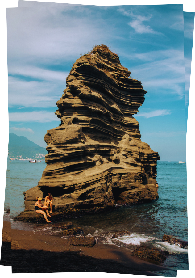 Vertical postcard of two bathers sitting on the edge of a large rock formation on a beach